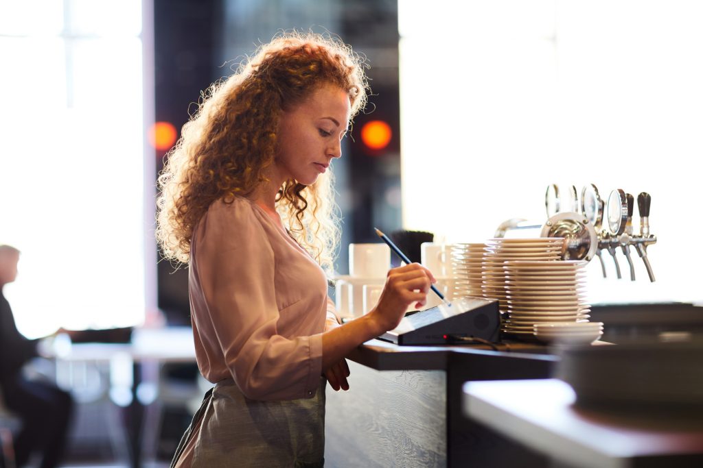 Waitress standing at bar counter and using pencil while adding order in restaurant POS system.